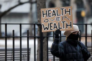 A bundled-up protester holds a cardboard sign reading "Health Over Wealth" during a public demonstration, symbolizing concerns over healthcare priorities amidst scrutiny of UnitedHealth's Medicare billing practices.