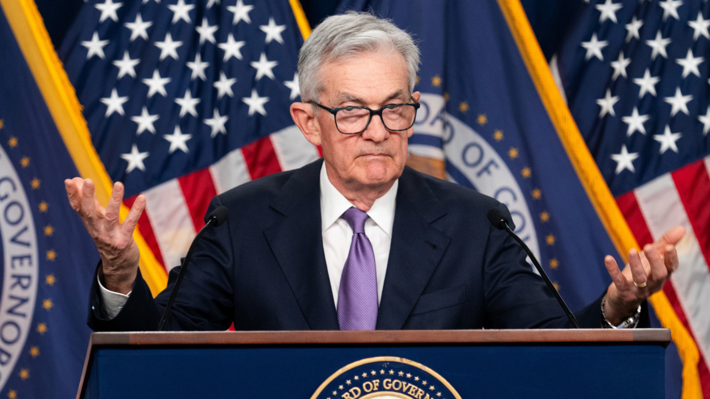 Federal Reserve official speaks at a press conference with U.S. flags in the background, addressing economic and monetary policy updates.