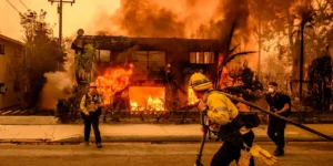 “Firefighters battle intense flames engulfing a residential building during Los Angeles wildfires, causing $65 billion in economic losses.